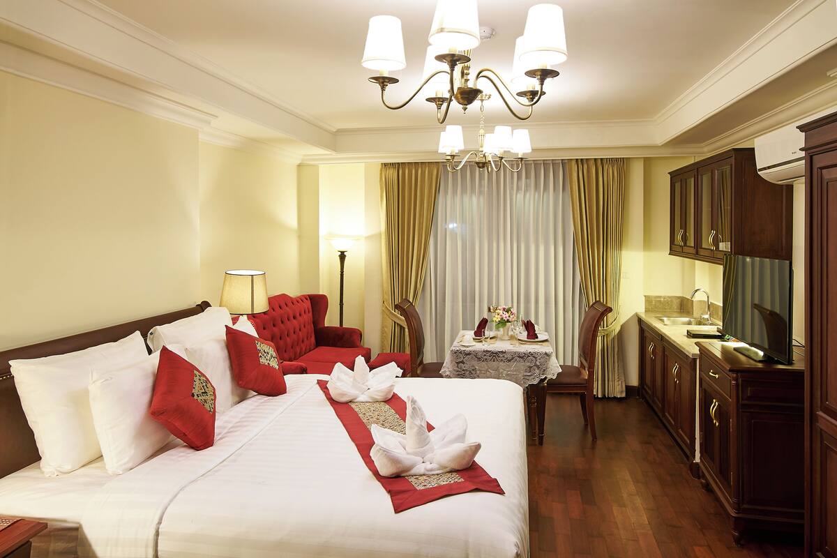 A hotel room with a large bed, a desk, a chair, a small table with a lamp, and a television.