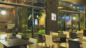 An outdoor dining area with tables and chairs, surrounded by glass walls and greenery. - High Livin Apartment Baros (Cimahi)