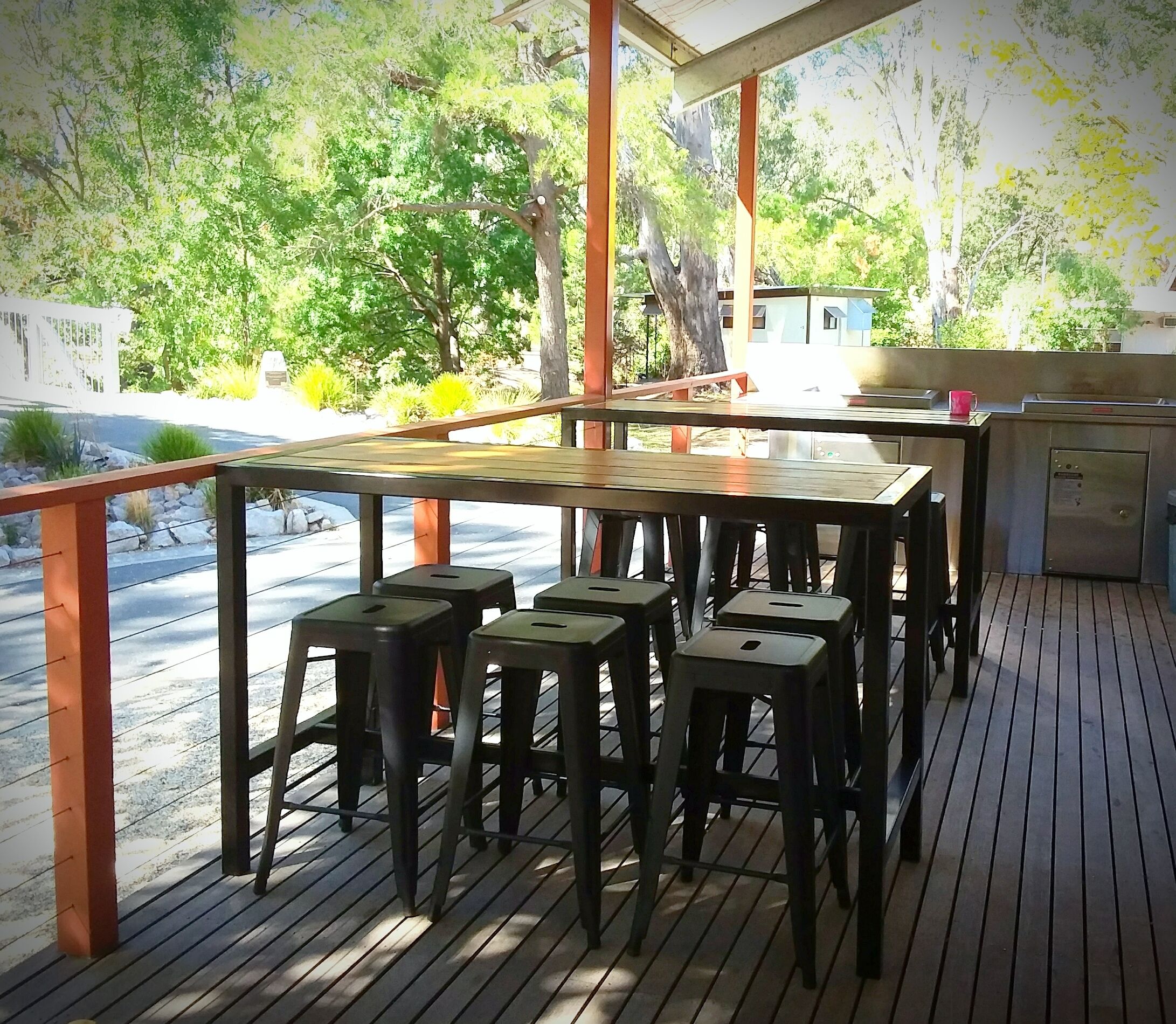 An outdoor deck with wooden tables and stools, a metal railing, and a view of trees and buildings in the background.