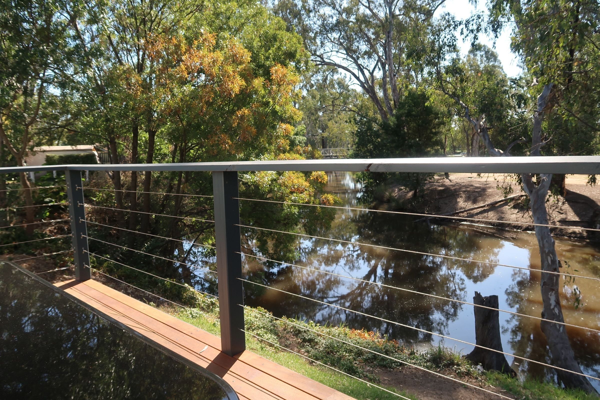 A wooden bridge over a calm river with a metal railing, surrounded by trees and greenery.
