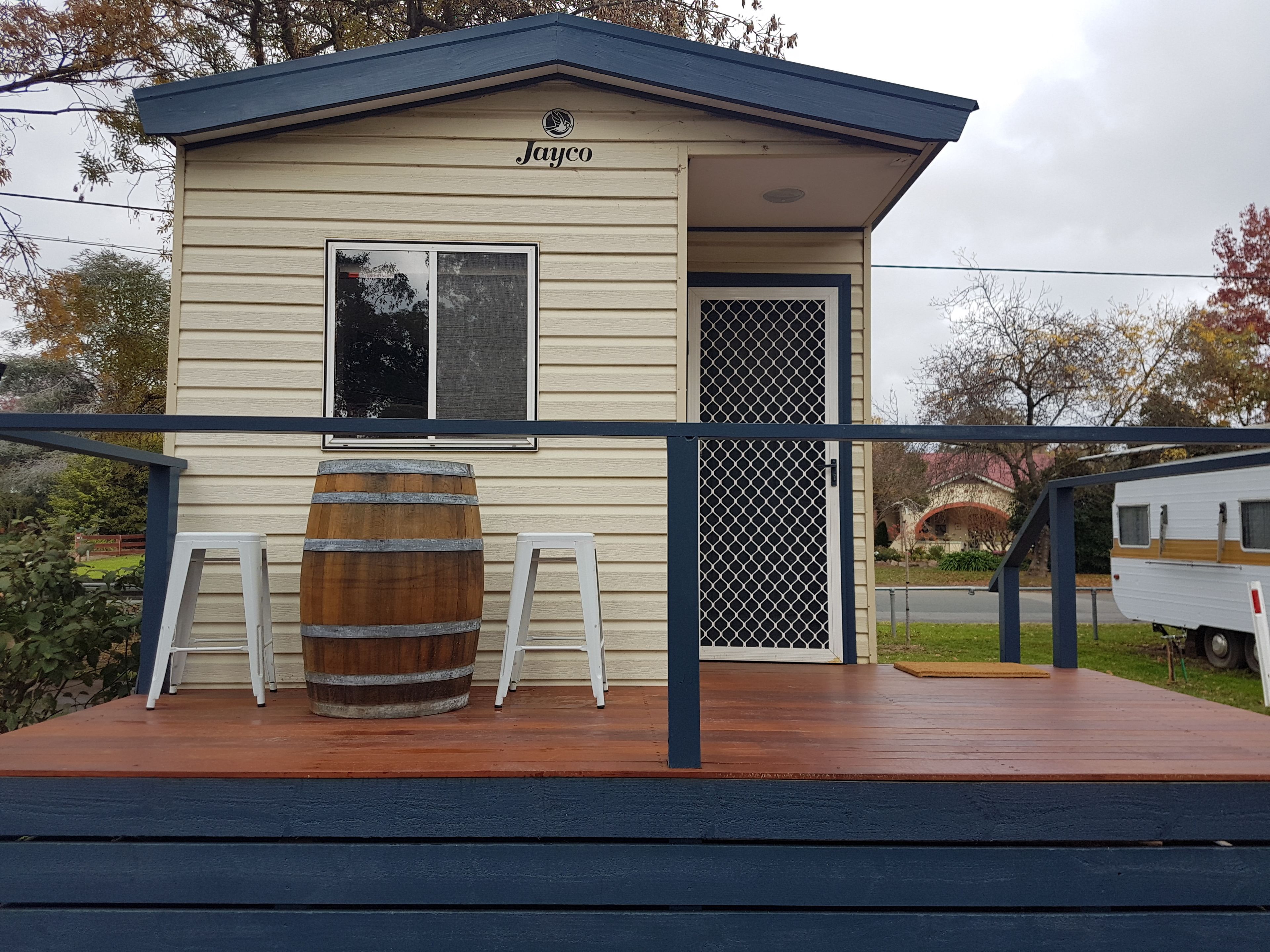 A small, single-story building with a dark blue roof and beige siding, labeled "Jayco." It features a wooden barrel, a white folding chair, and a metal gate.