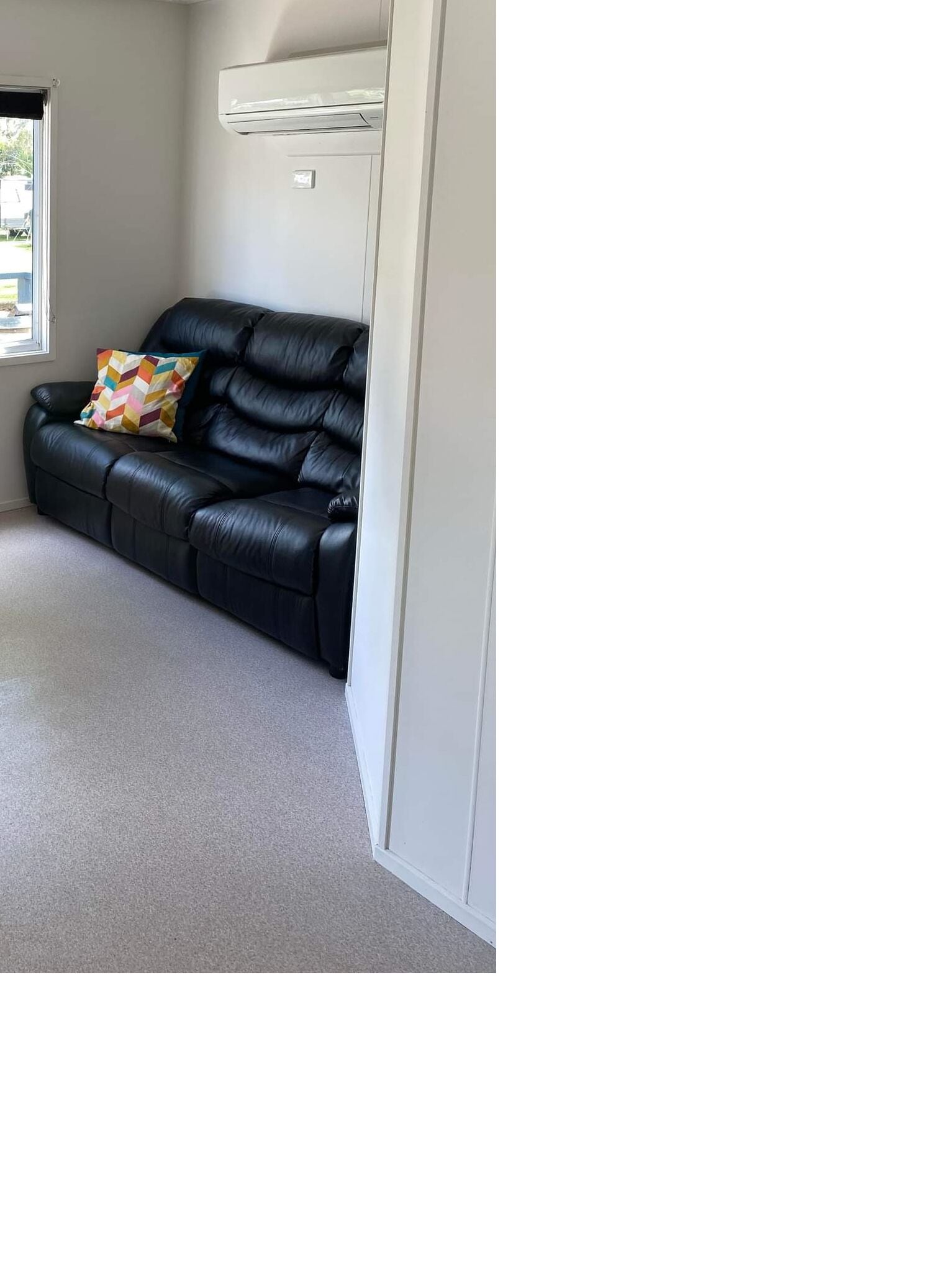 A room with a black leather sofa, a colorful cushion, a white door, and a carpeted floor.
