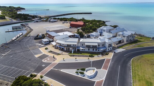 A modern hotel with a waterfront view, featuring a central courtyard, a parking lot, and a marina with boats.
