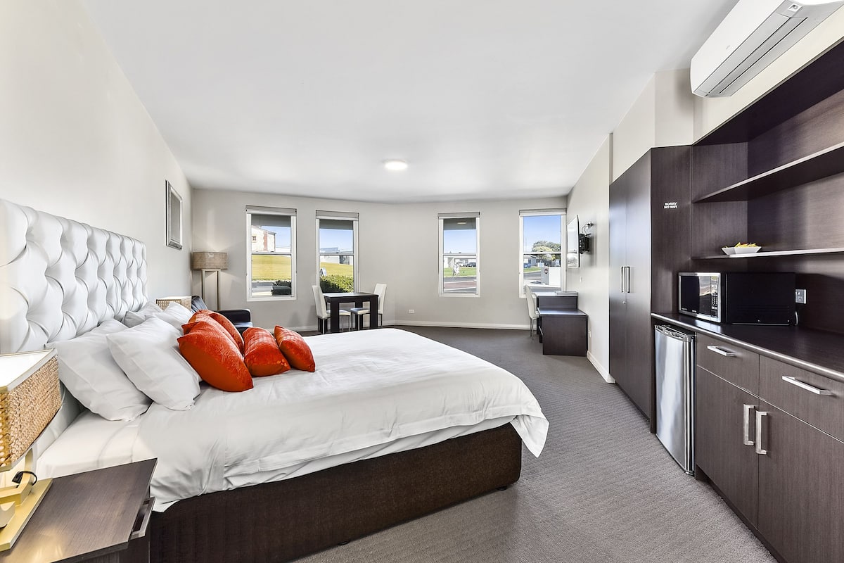 A modern hotel room with a large bed, a desk, and a kitchenette.