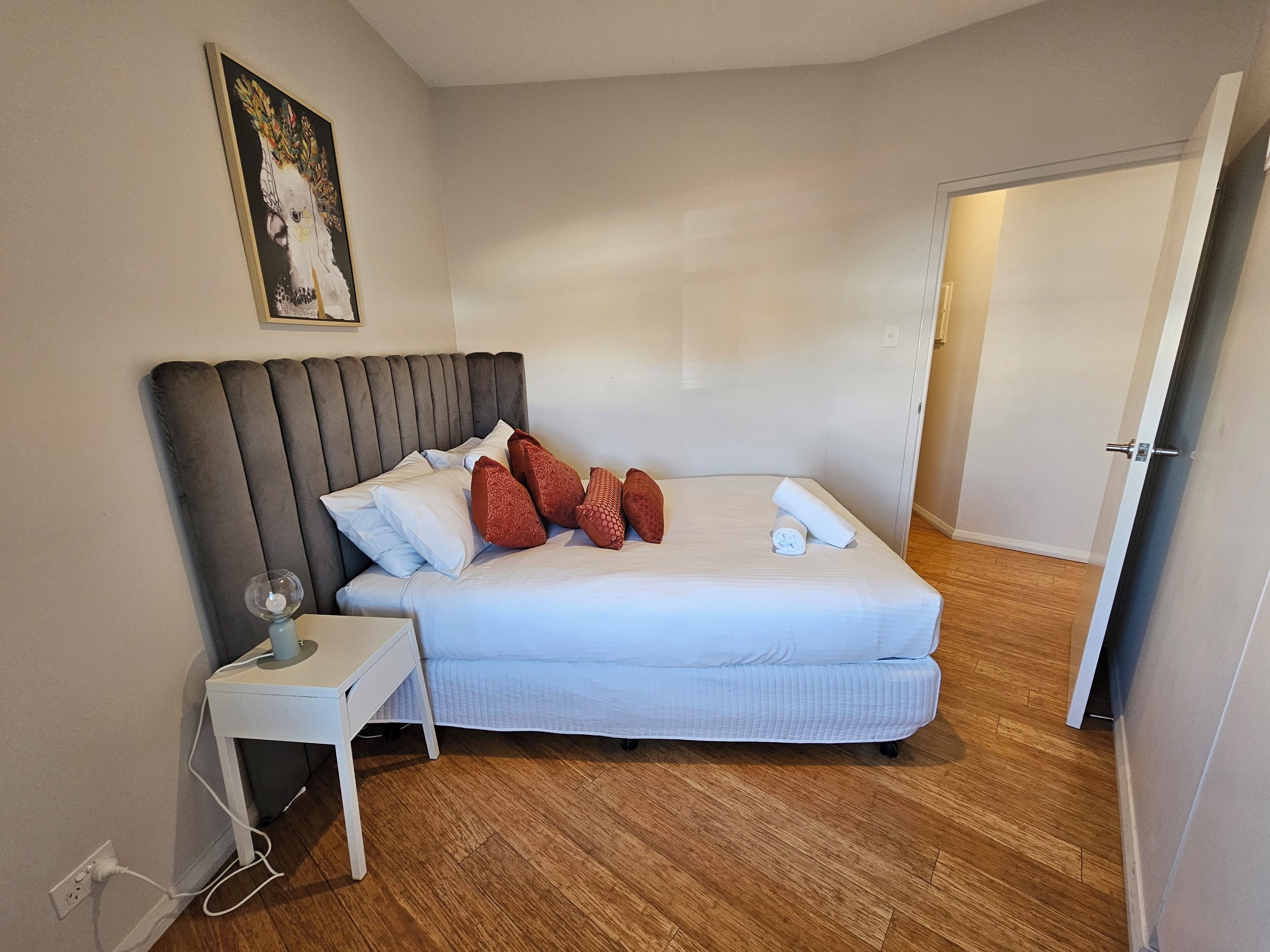 A neatly made bed with a grey upholstered headboard, a white bedside table with a lamp, and a painting on the wall.