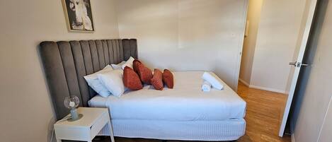 A neatly made bed with a grey upholstered headboard, a white bedside table with a lamp, and a painting on the wall.