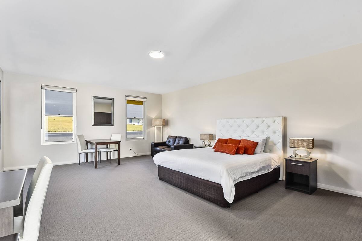 A modern hotel room with a large bed, a desk, and a chair. There are two windows and a view of the outside.