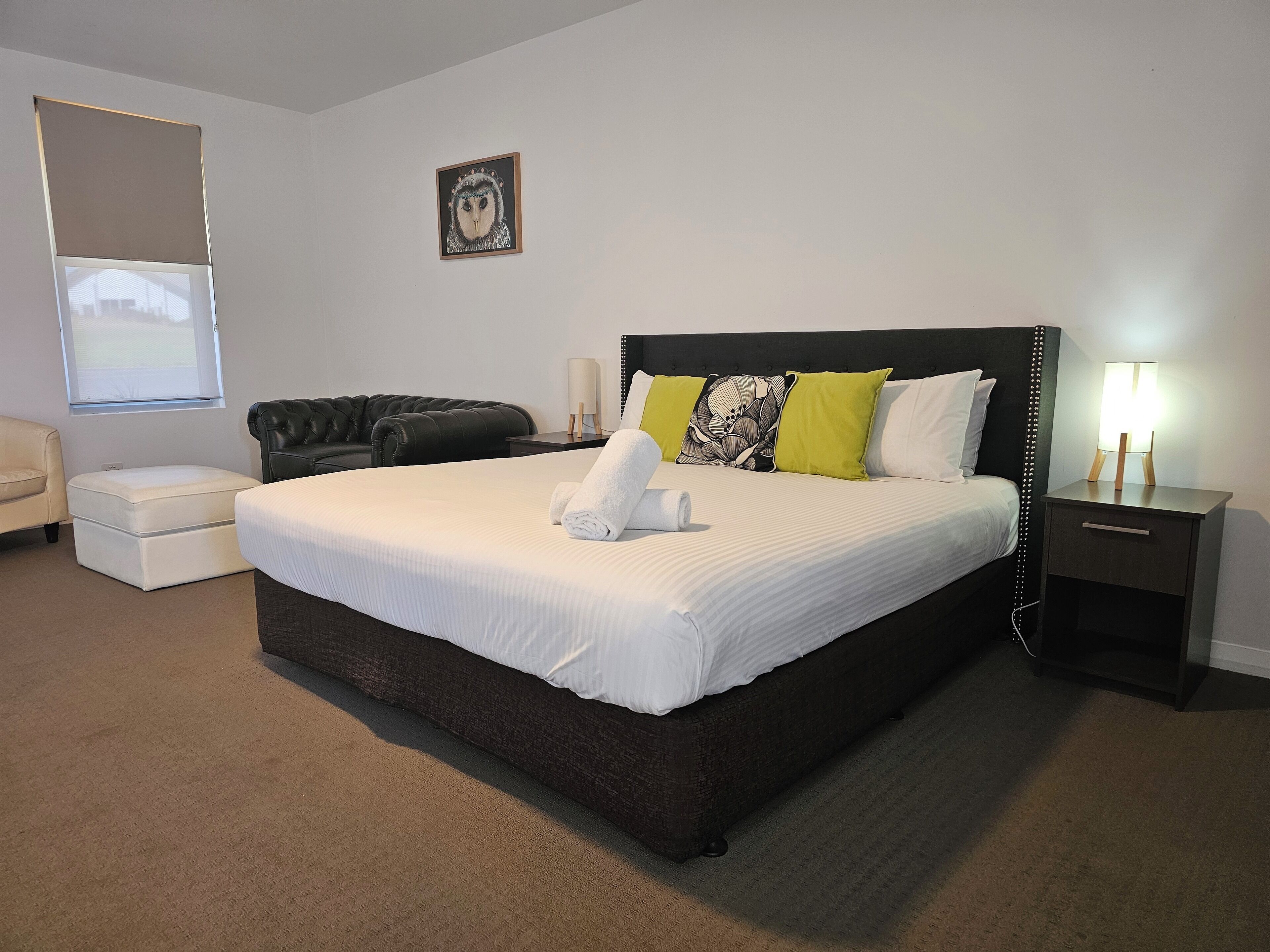 A neatly arranged bedroom with a large bed, a black leather sofa, a small white ottoman, and a nightstand with a lamp.
