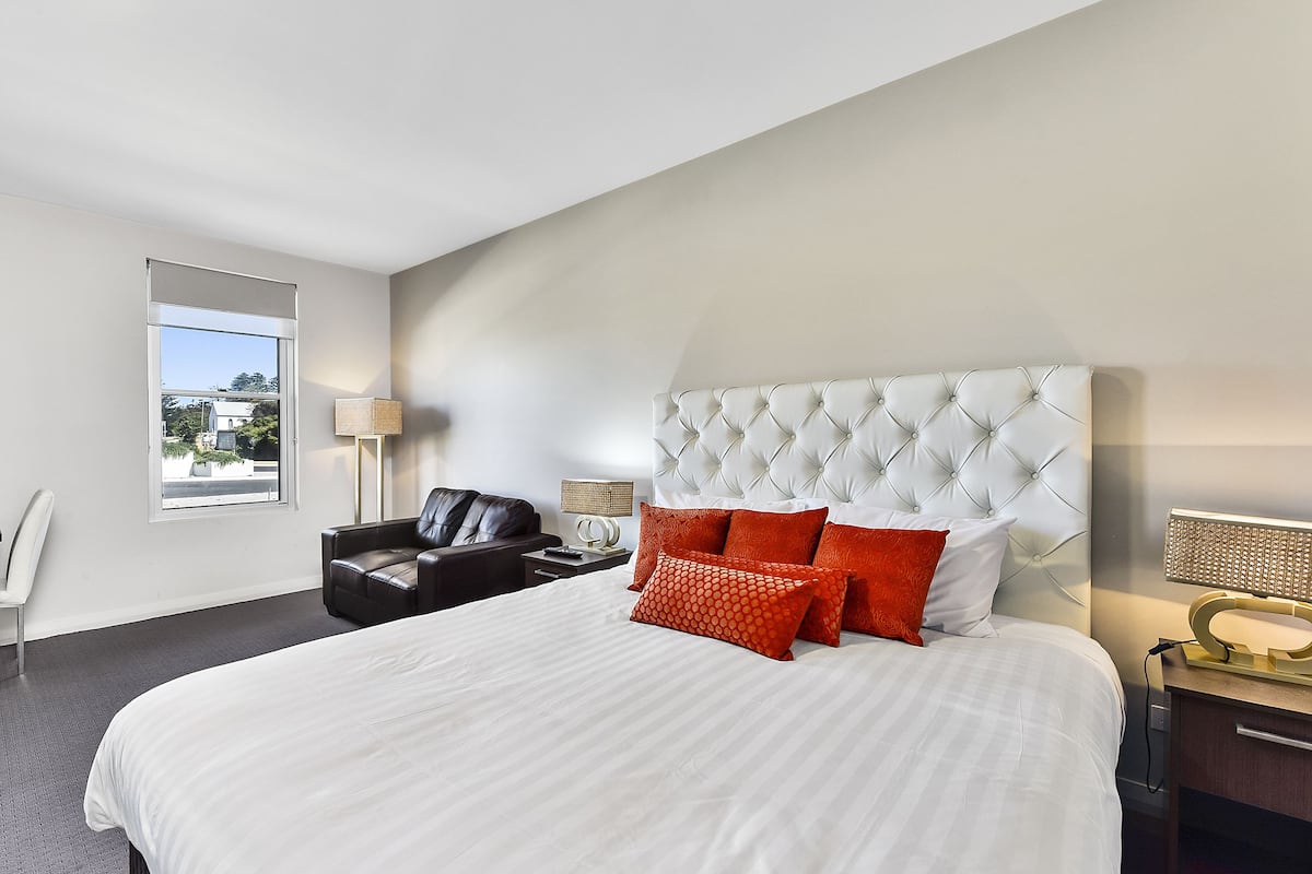 A hotel room with a large bed, a leather chair, a nightstand, and a window with a view of buildings.