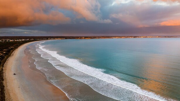 An aerial view of a beach with a curved shoreline, waves breaking onto the sand, and a distant town with lights. - Robe Marina Accommodation (Robe)