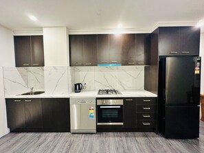 A modern kitchen with dark wood cabinets, a stainless steel dishwasher, a black refrigerator, a gas stove, and a built-in oven. - Exodus Dandenong Apartment Hotel (Melbourne)