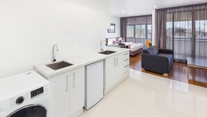 A modern kitchen with a dishwasher, a sink, and a compact washer. There is a dining area with a sofa and a table. The room has large windows with curtains. - Exodus Dandenong Apartment Hotel (Melbourne)