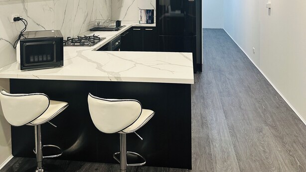 A modern kitchen with dark cabinets, a marble backsplash, a black refrigerator, and a central island with two white bar stools. - Exodus Dandenong Apartment Hotel (Melbourne)