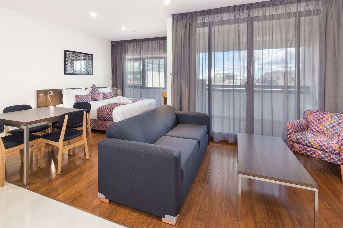 A modern hotel room with a sofa, a dining table with chairs, a bed with patterned pillows, and large windows with sheer curtains.