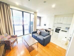 A modern living room with a sofa, a patterned armchair, a coffee table, and a sliding glass door leading to a balcony. - Exodus Dandenong Apartment Hotel (Melbourne)