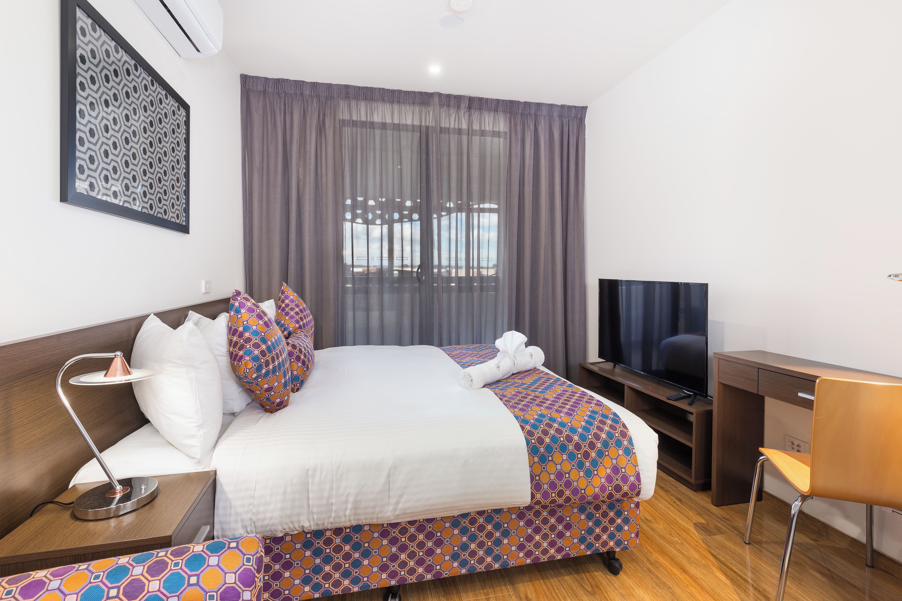 Habitación de hotel con cama, escritorio, silla, televisor y ventana con cortinas.