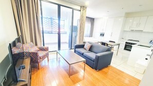 A modern living room with a sofa, coffee table, and patterned chairs. A kitchen with white cabinets and a dining area is visible in the background. - Exodus Dandenong Apartment Hotel (Melbourne)