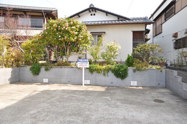 Un edificio residencial de estilo arquitectónico japonés tradicional, con tejado de tejas y elementos de madera. Cuenta con un pequeño jardín con diversas plantas y un cartel que indica un espacio de estacionamiento.