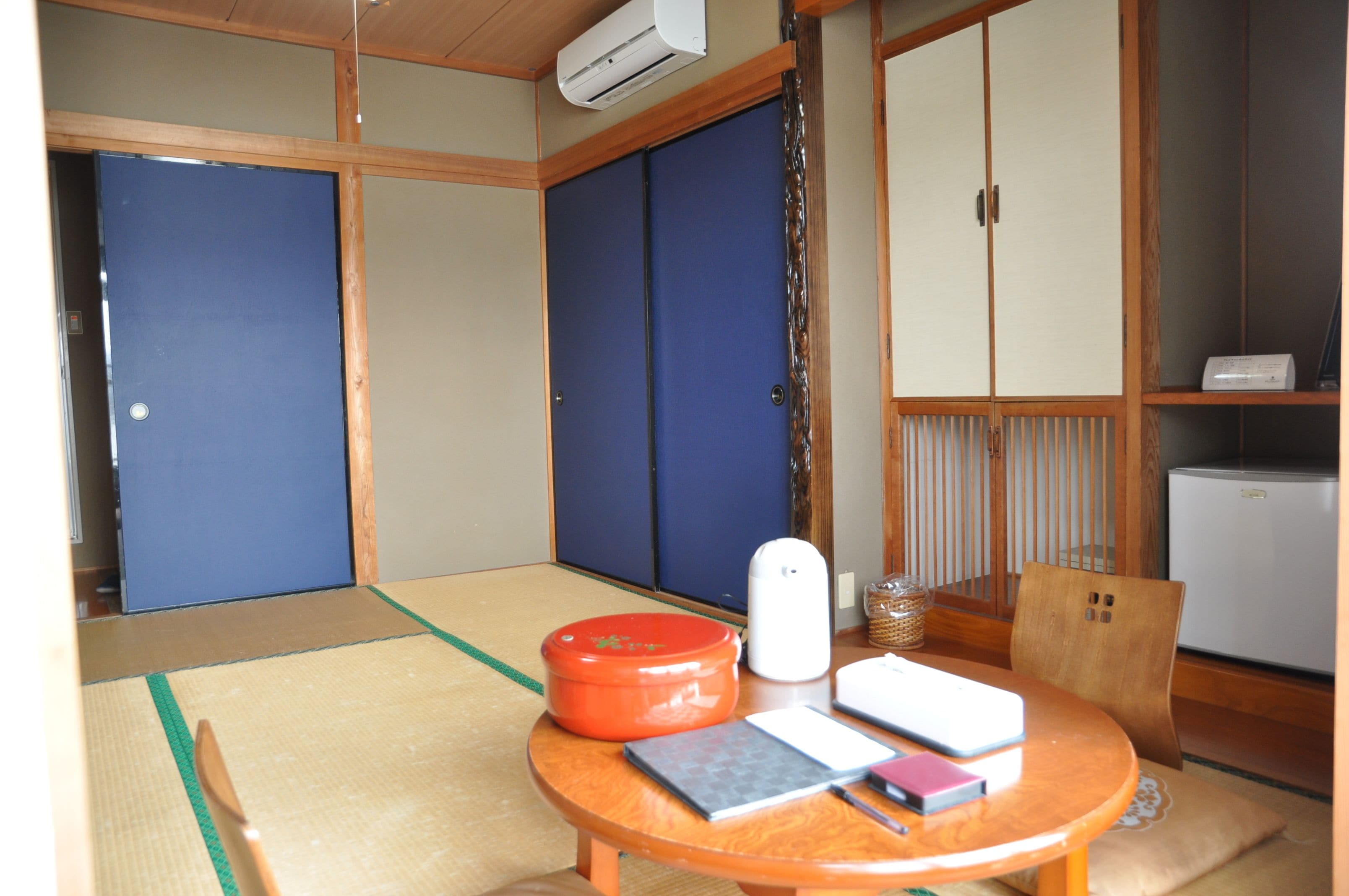 Una habitación japonesa tradicional con suelo de tatami, una mesa de madera baja con libros y un recipiente rojo, y un aire acondicionado blanco.