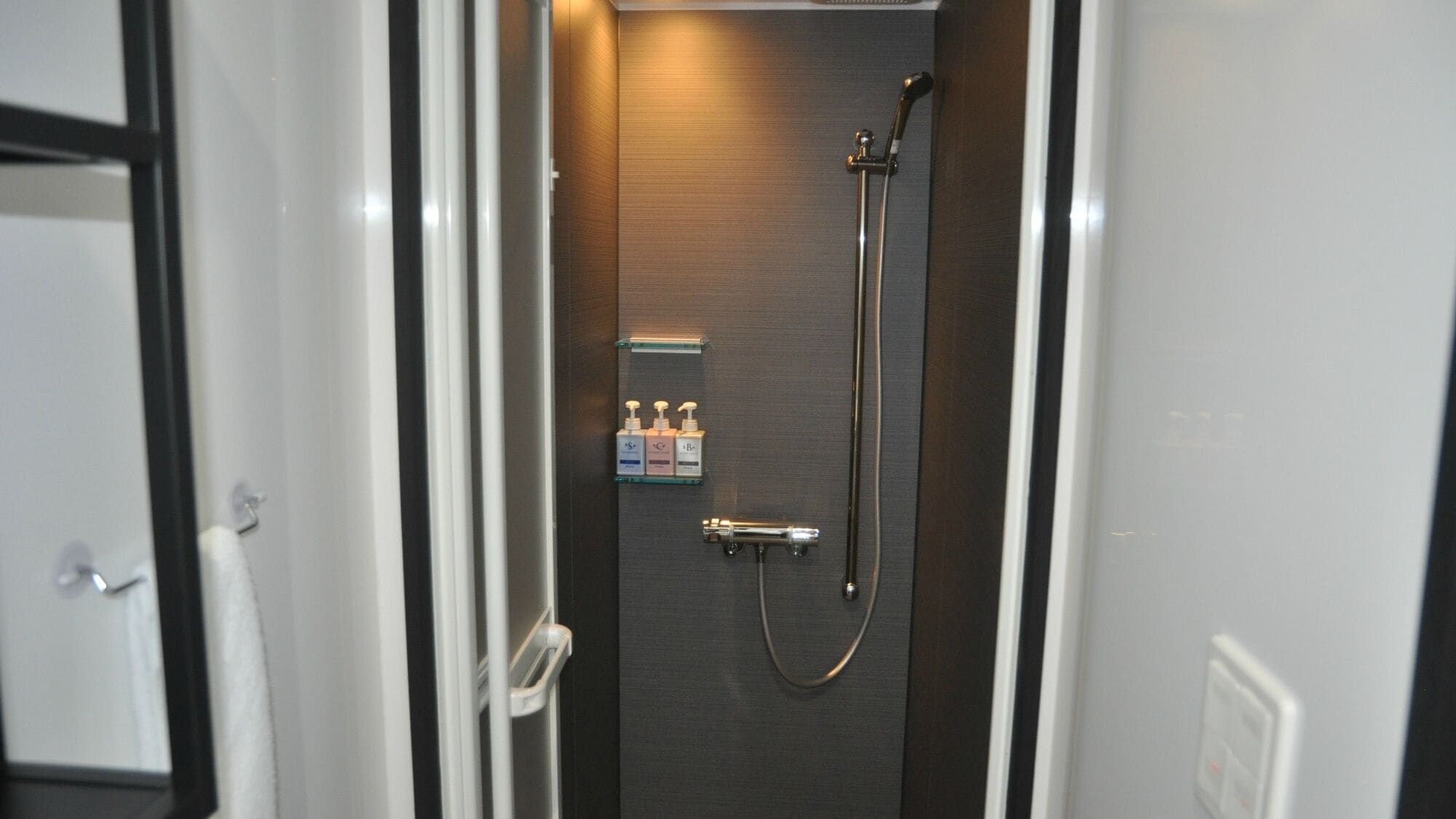 A narrow bathroom with a shower, a towel rack, and a shelf with toiletries.