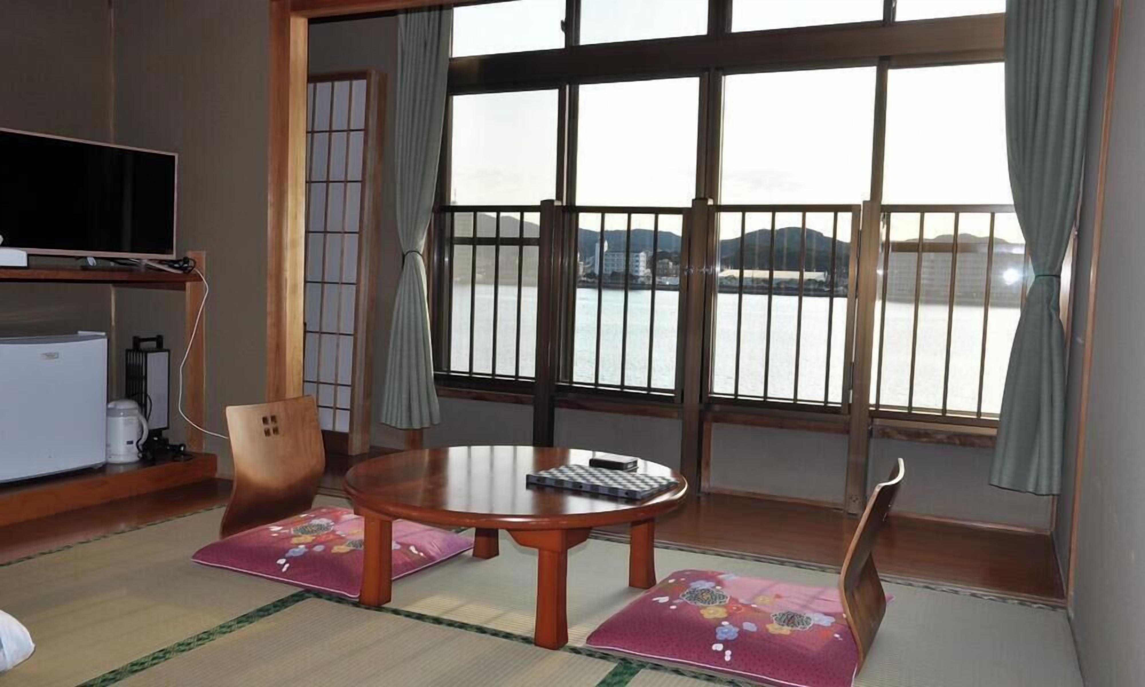 A room with tatami mats, a wooden table, a TV, and a view of a body of water through large windows.