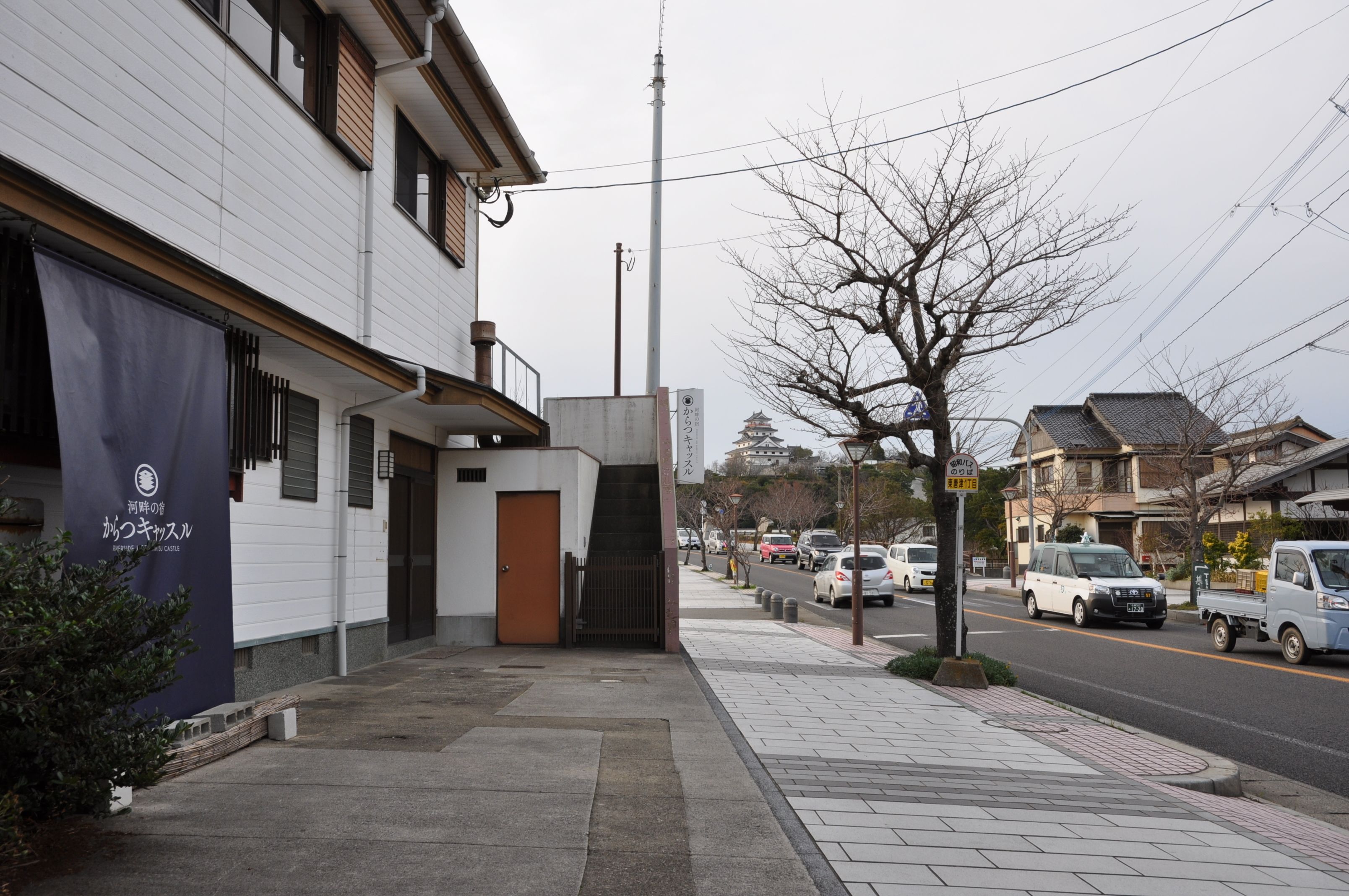 Ein Gebäude mit weißer Fassade, einer braunen Tür und einem blauen Banner mit japanischem Text. Auf der Straße sind Autos geparkt und ein Baum ohne Blätter.