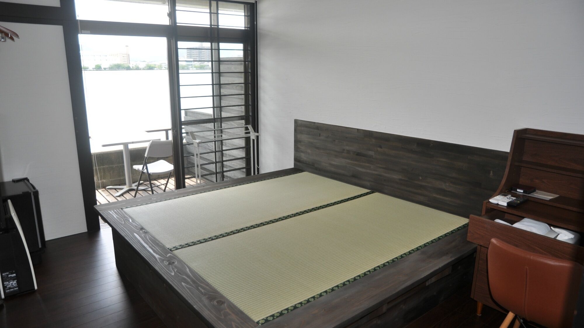 Quarto com cama, cadeira, escrivaninha e janela com vista para prédios.