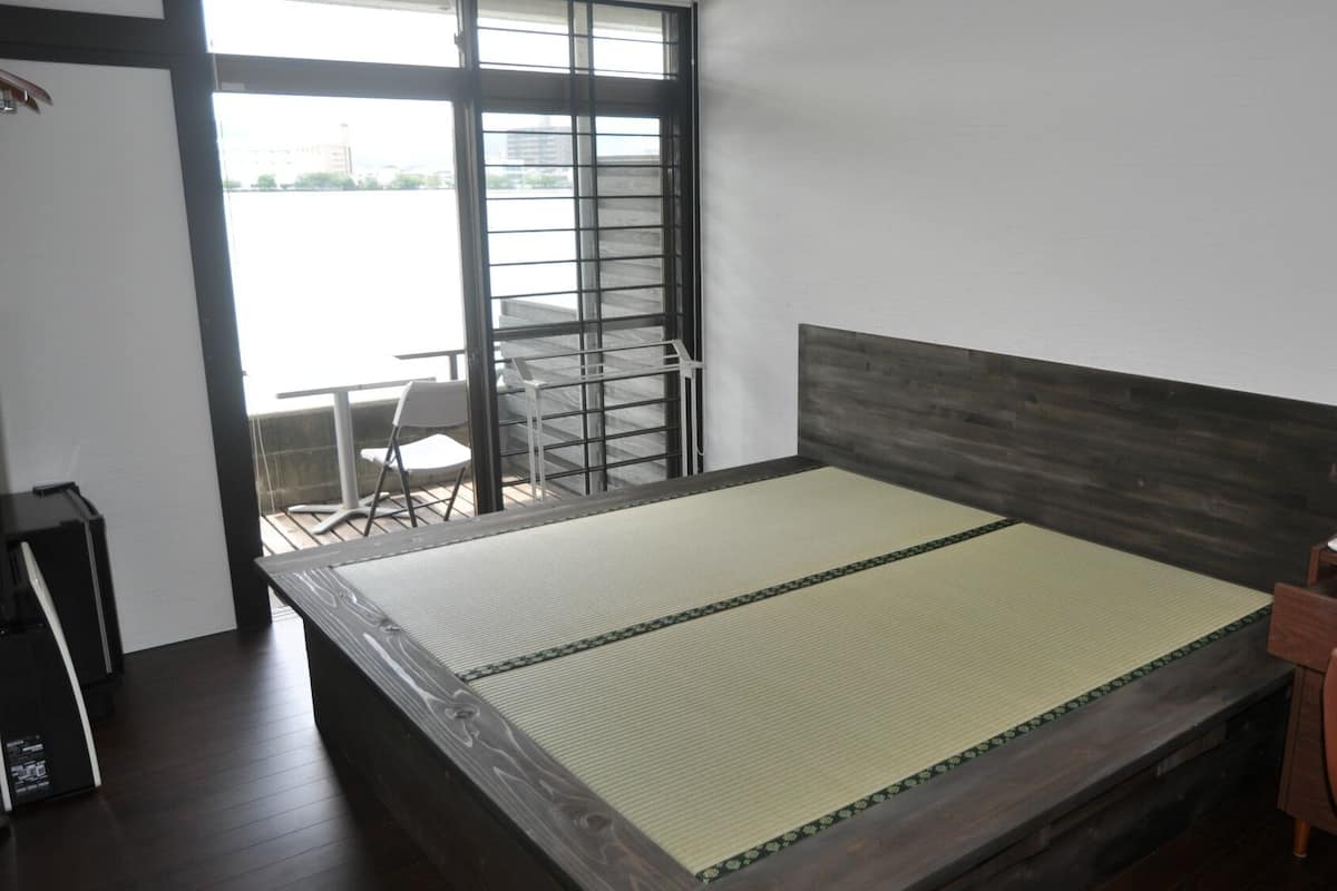 Quarto com cama, cadeira, escrivaninha e janela com vista para prédios.