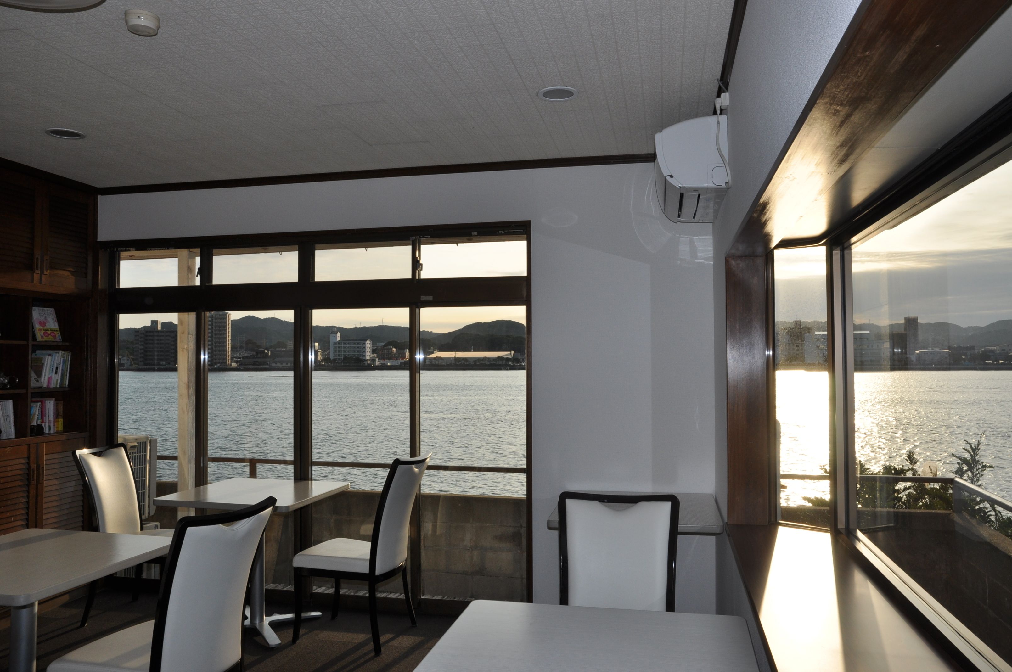 A dining area with tables and chairs, large windows with a view of a body of water and distant buildings.