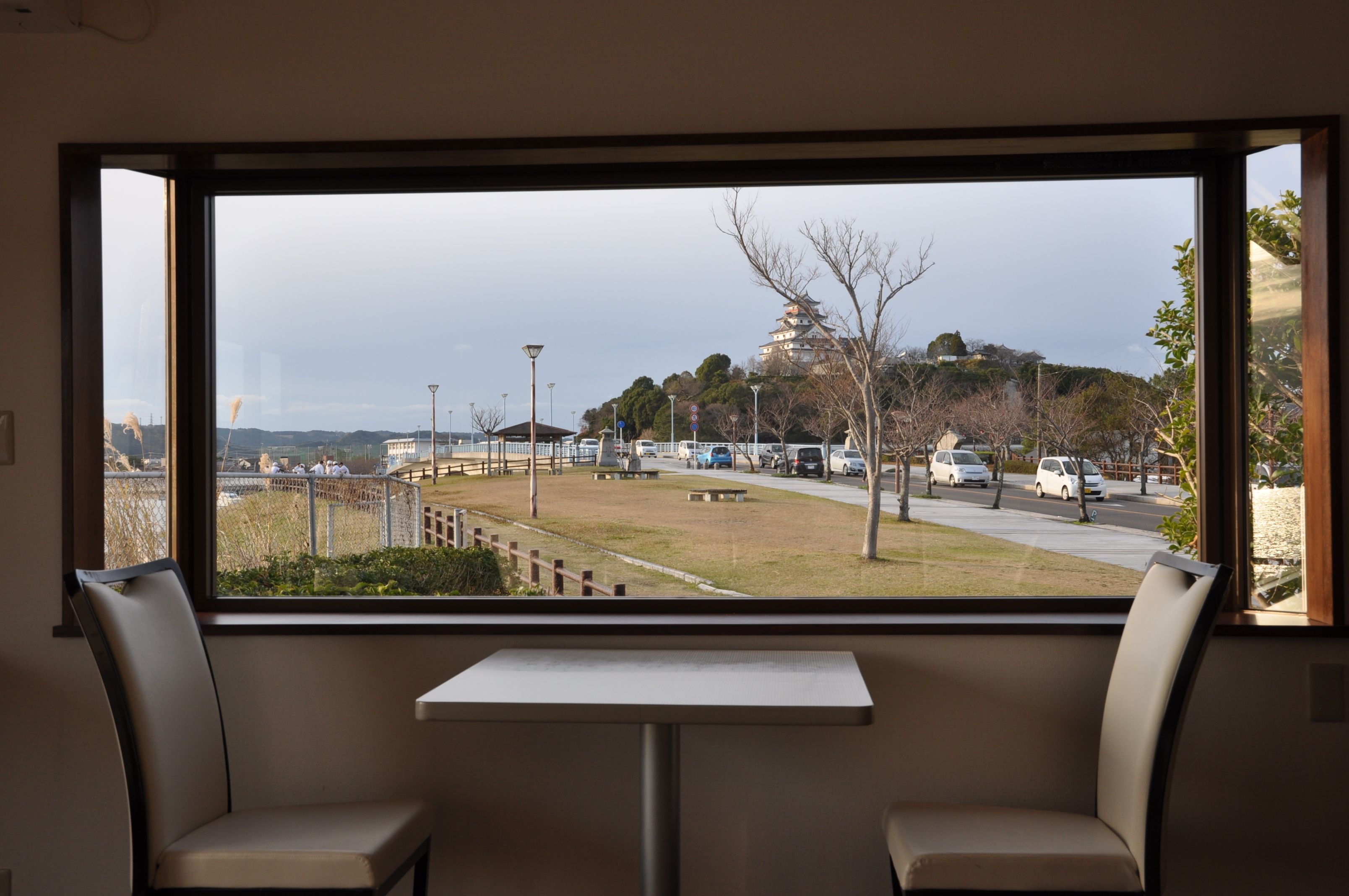 Vista desde una ventana de una habitación con dos sillas y una mesita. Se ve un estacionamiento con coches y un edificio con una estructura similar a una pagoda.