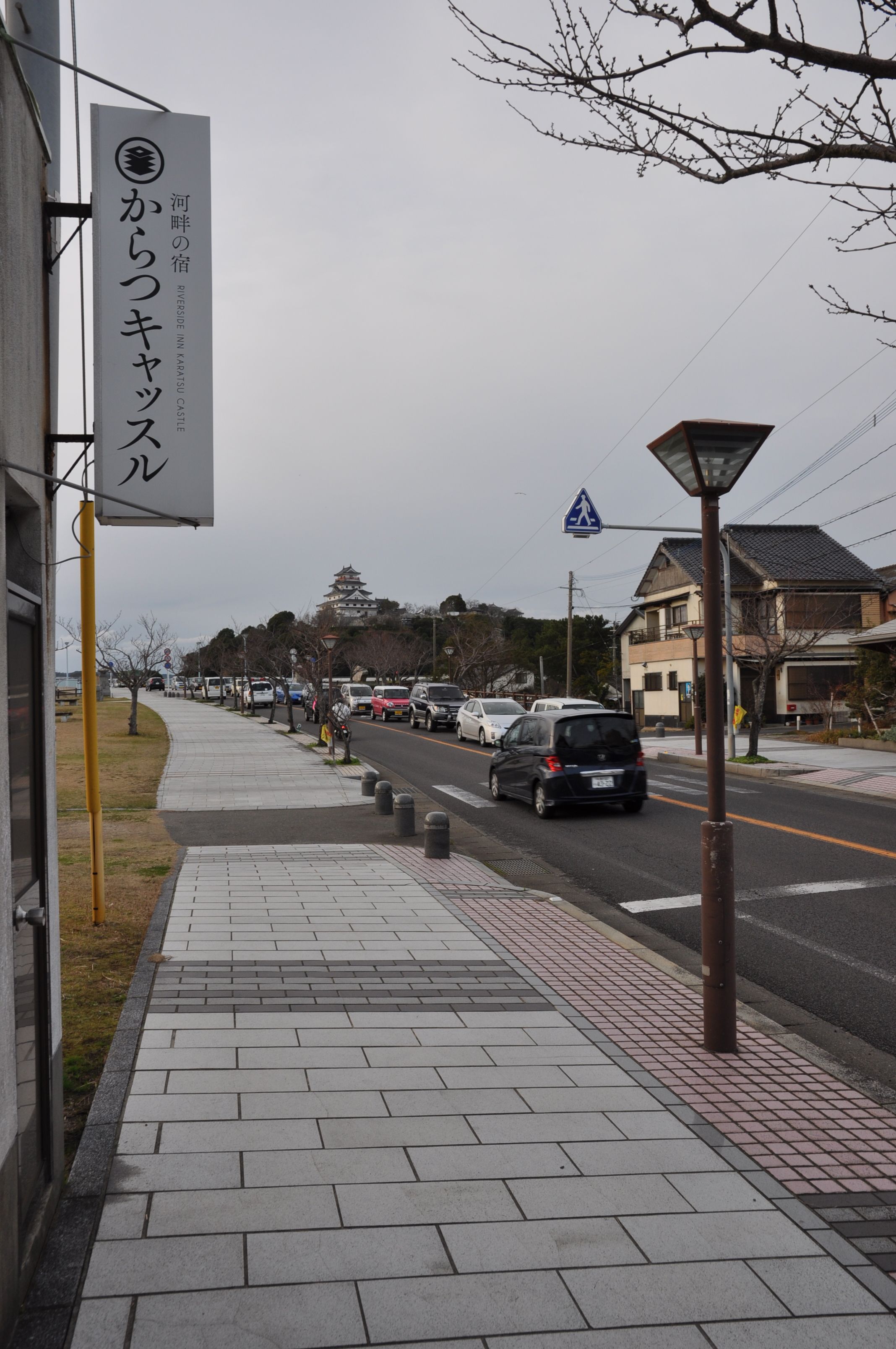 Une rue où les voitures sont garées le long du trottoir, un panneau indiquant un passage pour piétons et un bâtiment avec une enseigne en japonais.