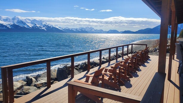 A wooden deck with chairs facing a rocky shoreline and a view of a calm sea with mountains in the background.