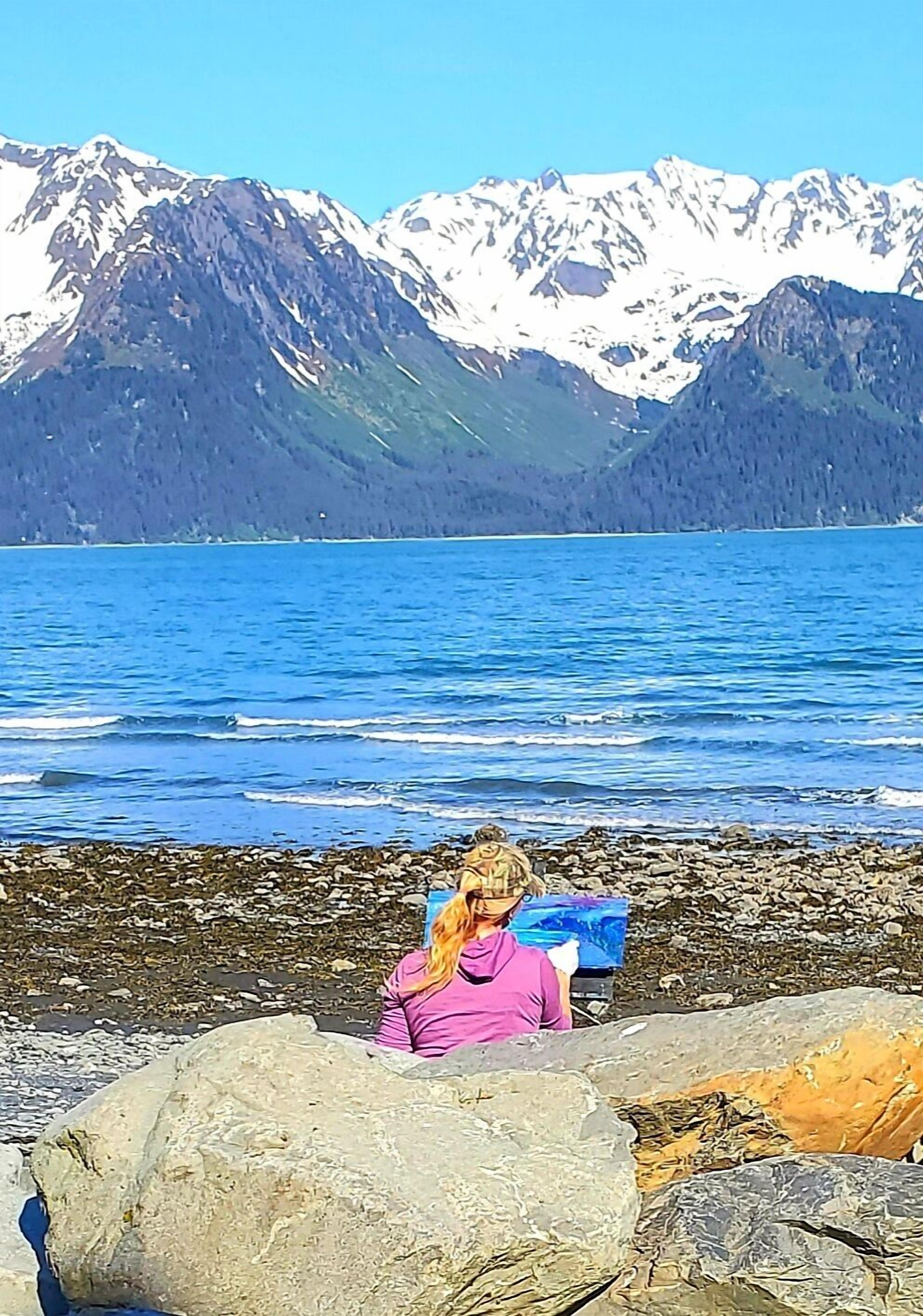 A person sitting on a rocky shore with a painting, facing a lake with mountains in the background.