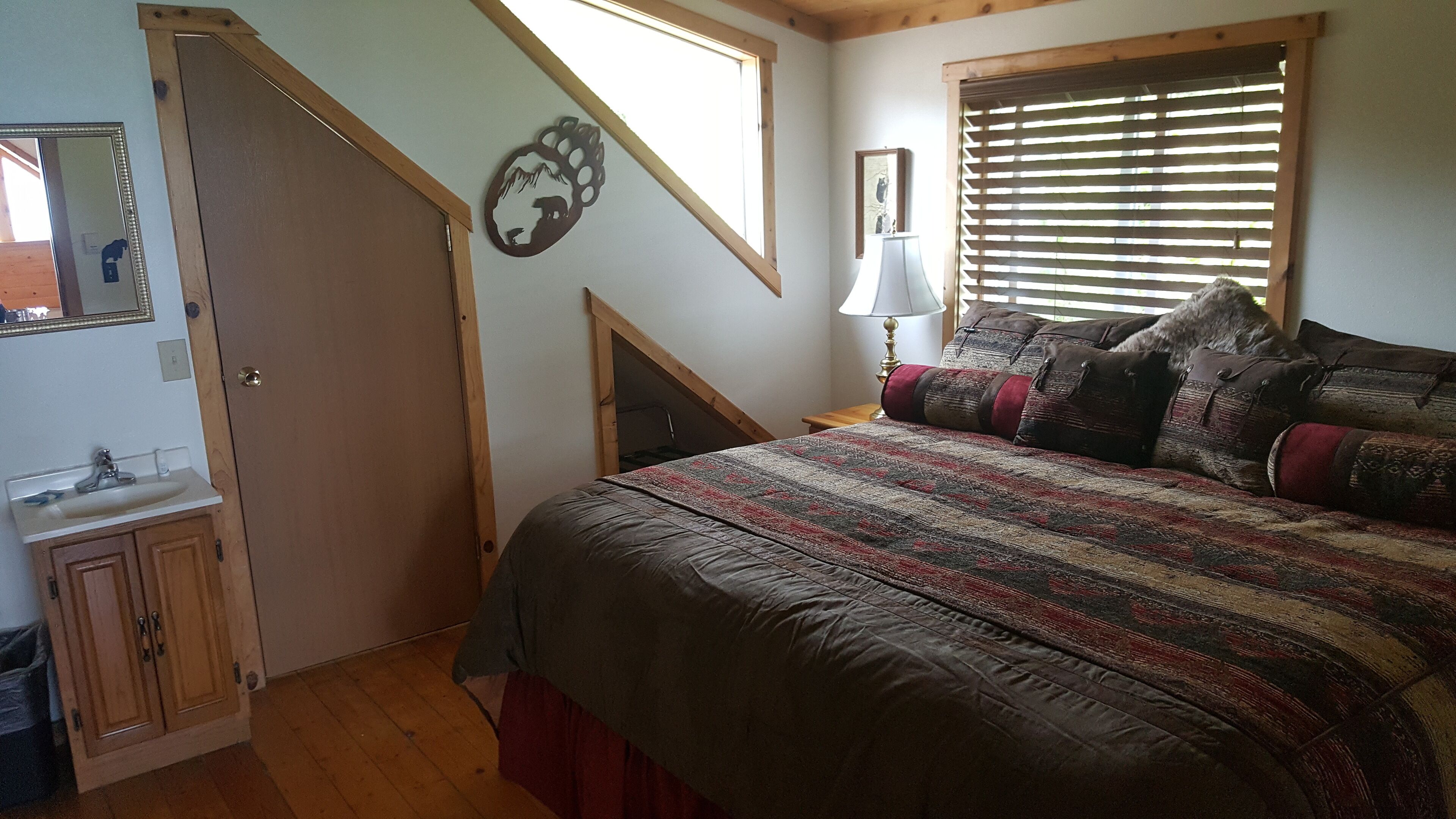 Un dormitorio con cama, mesita de noche, lámpara, ventana con persianas, puerta de madera, espejo y decoración en la pared.