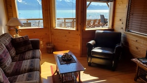 A cozy room with a wooden interior, a sofa, a chair, and a table with a decorative item. Large windows offer a view of the sea and mountains.