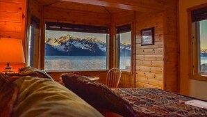 A cabin room with a bed, a chair, and a table. It has windows with a view of mountains and water.