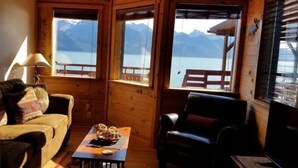 A cozy cabin room with a wooden interior, a sofa, a chair, and a table with a plate of food. Large windows offer a view of mountains and a lake.