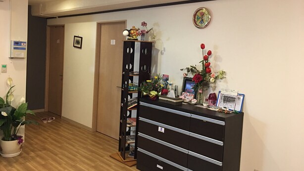 A reception area with a wooden floor, a black filing cabinet with drawers, a clock on the wall, and a potted plant. - Women's Guest House Fukutomi (Naha)