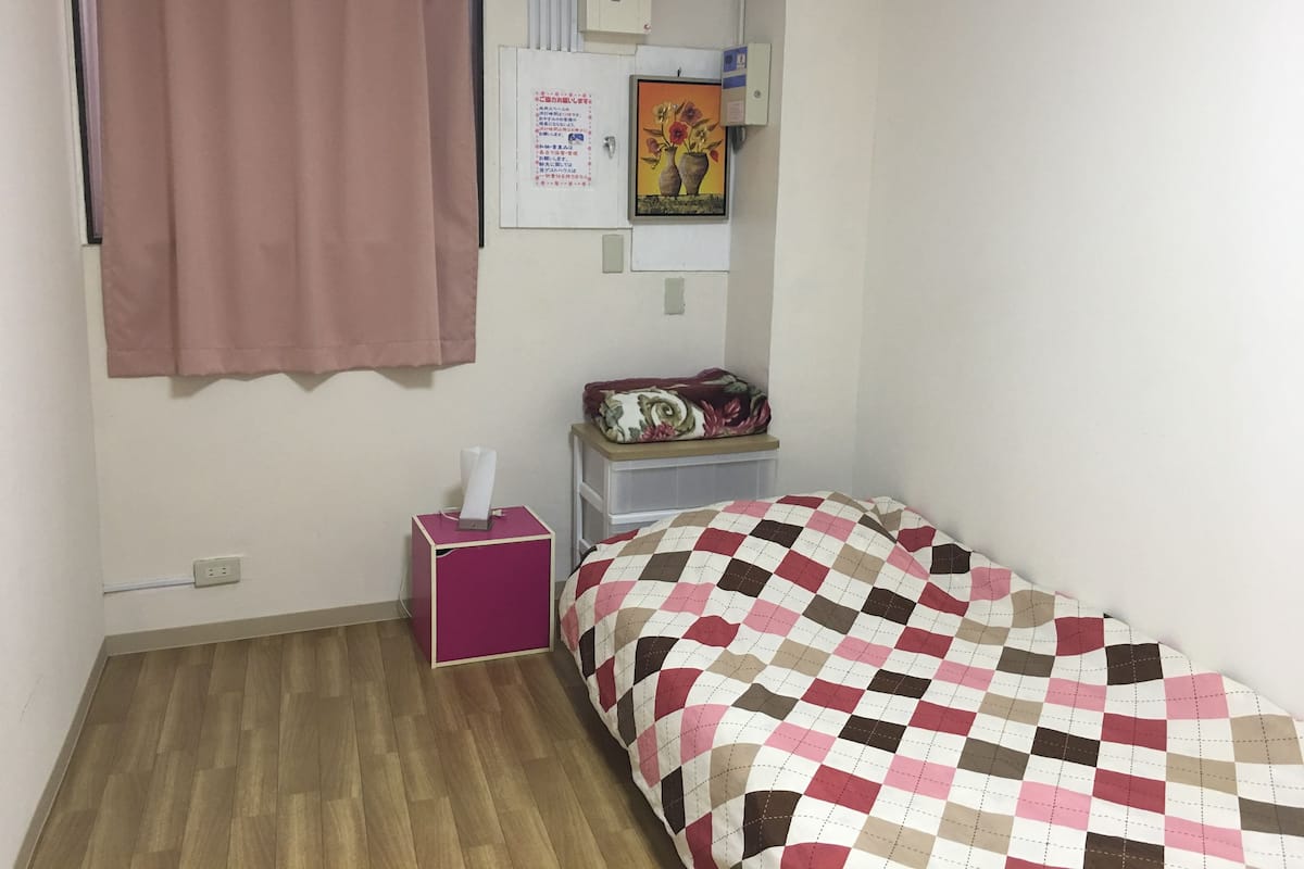 Ein kleines, ordentlich eingerichtetes Schlafzimmer mit einem Einzelbett, einem Nachttisch mit Taschentuchbox, einer Wand-Klimaanlage, einer Steckdose und einem Poster an der Wand.