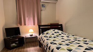 A small, neatly arranged bedroom with a bed, a bedside table with a lamp, a TV, and a shelf with items. - Women's Guest House Fukutomi (Naha)