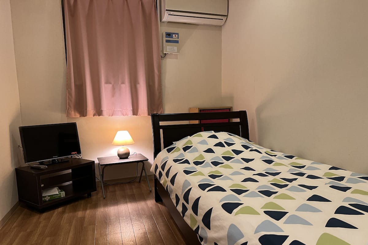 Ein kleines, ordentlich eingerichtetes Schlafzimmer mit Bett, Nachttisch mit Lampe, Fernseher und einem Regal mit Gegenständen.