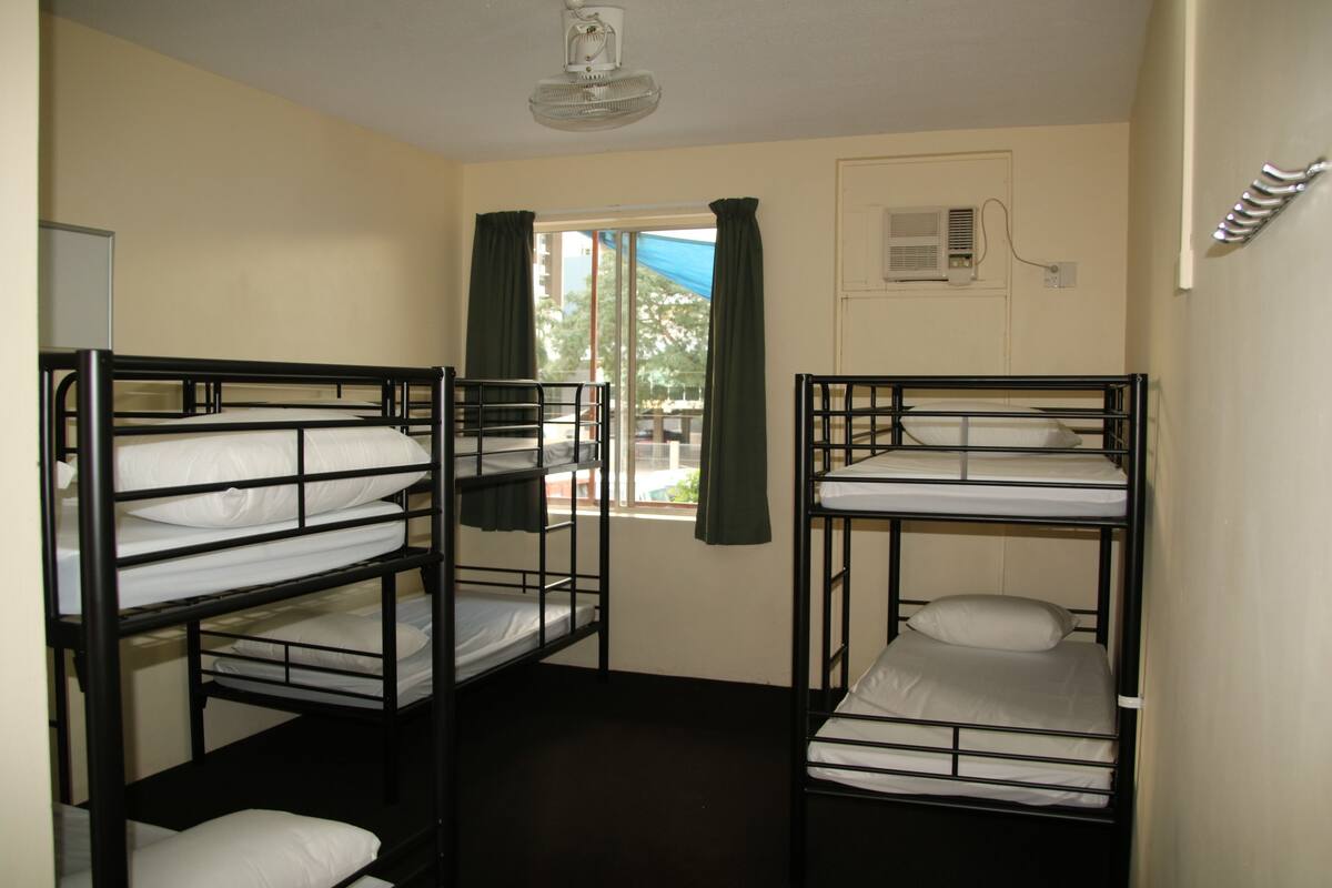 A dormitory room with bunk beds, a window with curtains, an air conditioning unit, and a ceiling fan.