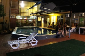An outdoor swimming pool area with lounge chairs and a group of people sitting around a table. - Crocodilly Inn - Hostel (Darwin)