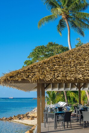 Un restaurant situé en bord de mer, doté d’une terrasse, d’un toit de chaume et offrant une vue sur l’océan.
