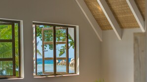 Un dormitorio con cama, ventilador de techo, ventana con vista a la playa y una planta sobre la mesita de noche.