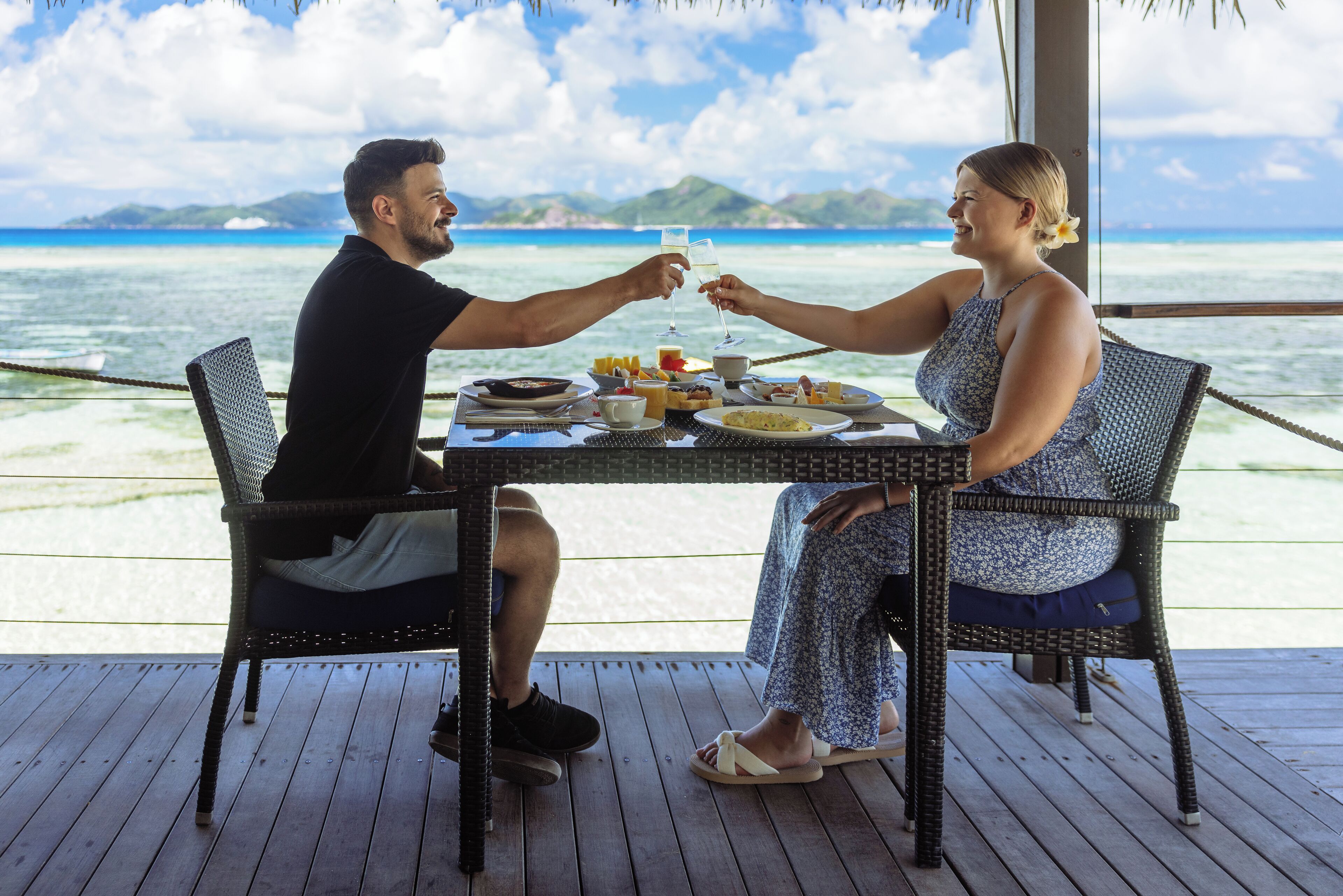 A couple toasting at a table with a view of the ocean and mountains.