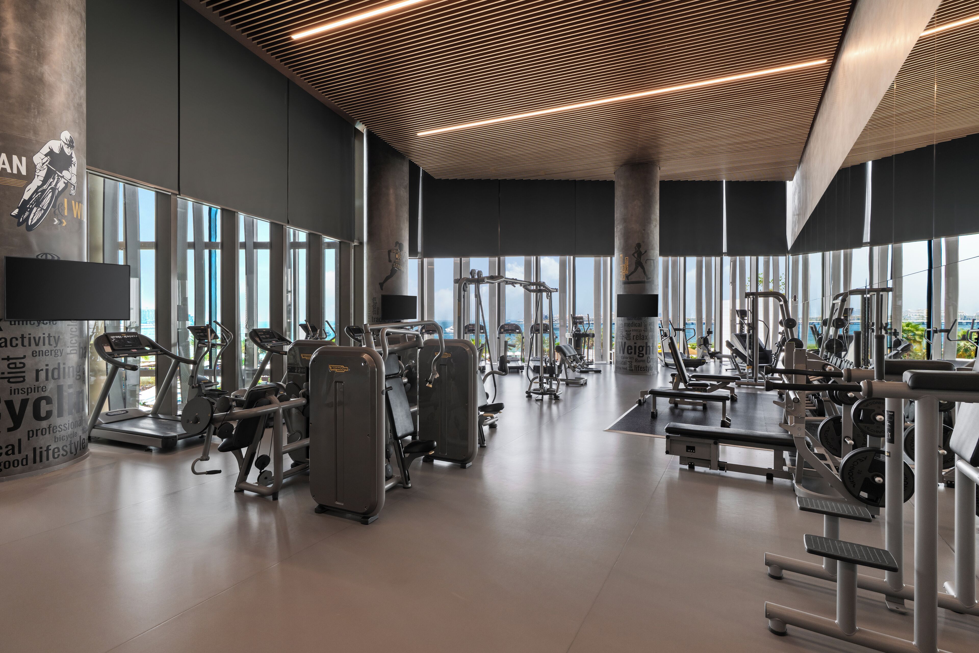 A modern gym with various exercise equipment, large windows, and motivational wall graphics.