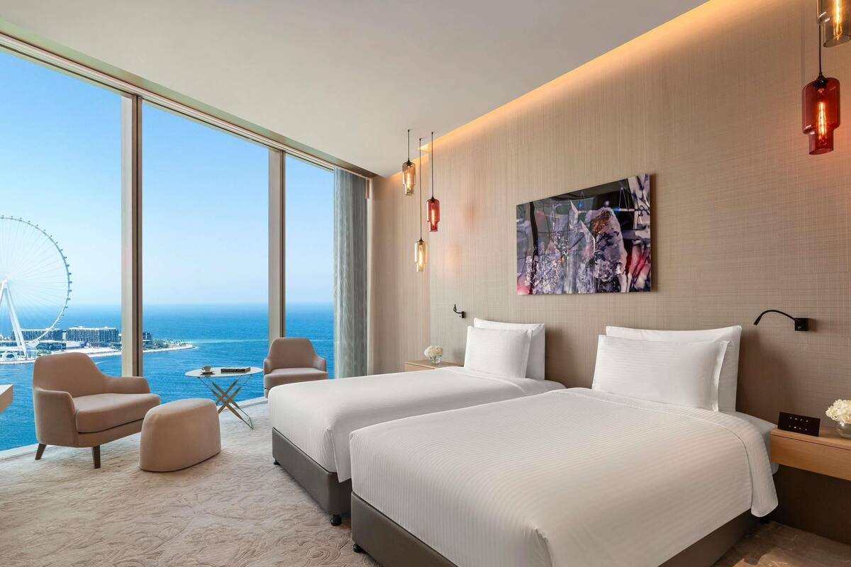 A hotel room with two beds, a large window with a view of the sea and a Ferris wheel, a small table, and a chair.