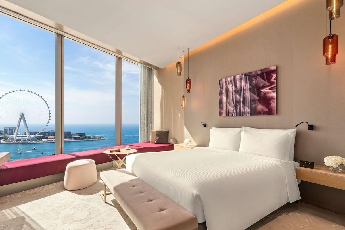 A modern hotel room with a large bed, a bench, a small table, and a view of a Ferris wheel across the water.