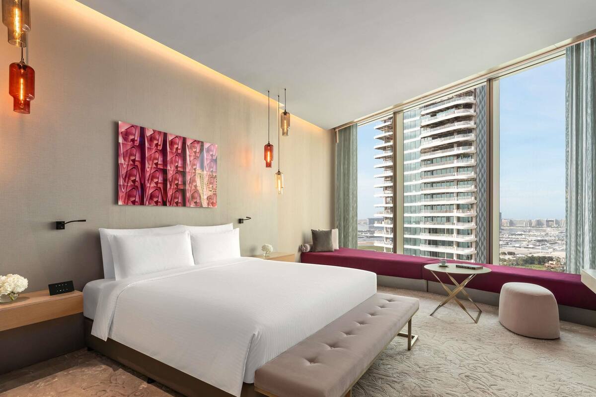 A modern hotel room with a large bed, a bench, a small table, and a view of high-rise buildings.